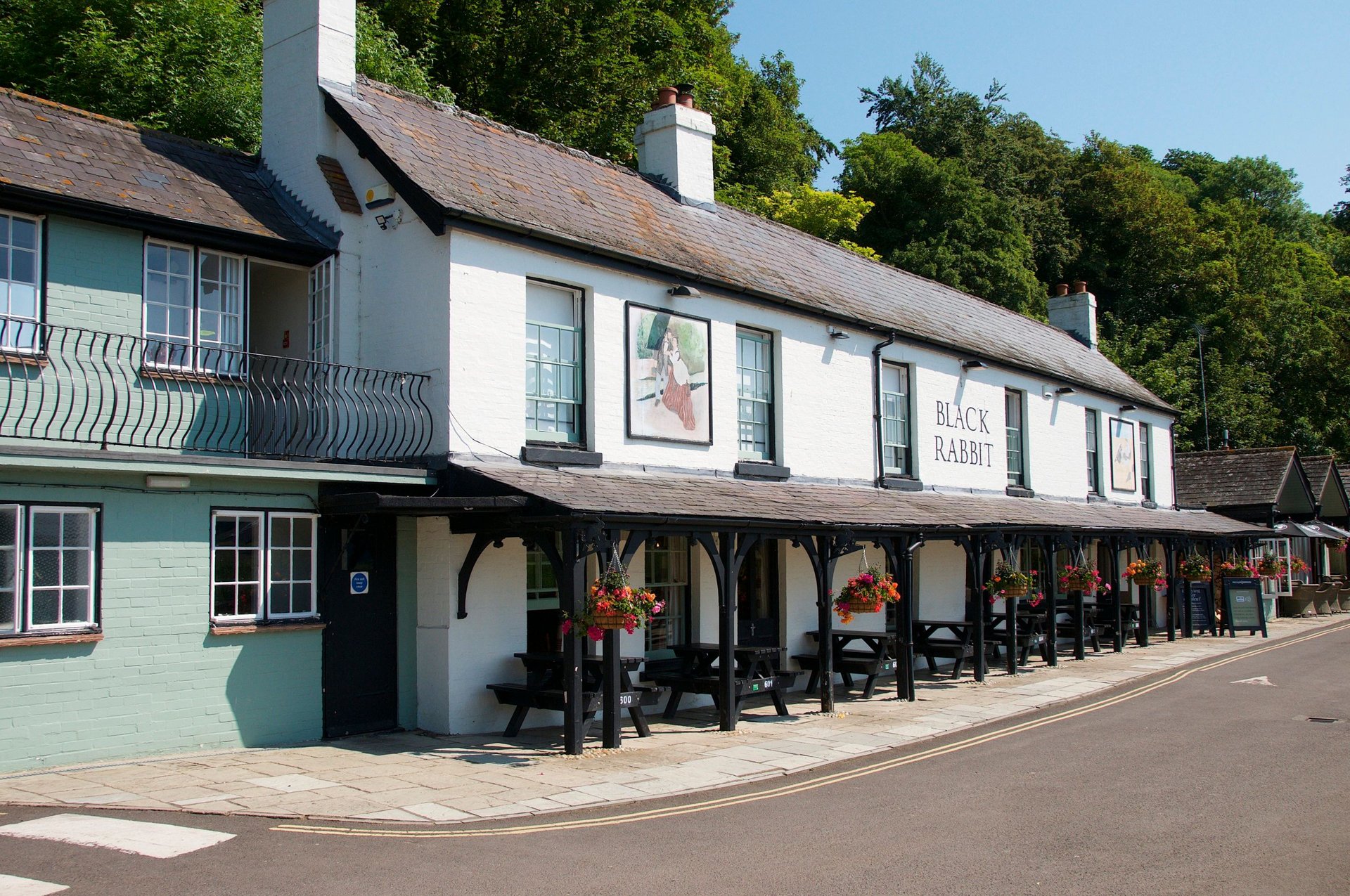 The white-painted front of The Black Rabbit at Offham, with its long veranda, hanging baskets and wooded slope behind